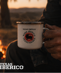 Person holding a 'Texas Iberico' mug by a campfire with sunset in the background