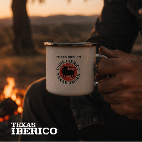 Person holding a 'Texas Iberico' mug by a campfire with sunset in the background