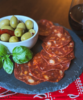 Platter of sliced salami with a bowl of olives on a wooden table with a glass of red wine.