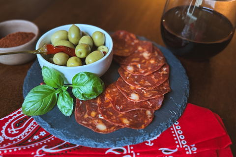Platter of sliced salami with a bowl of olives on a wooden table with a glass of red wine.