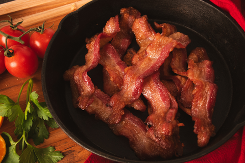 Cooked bacon strips in a black skillet on a wooden surface with tomatoes and herbs.